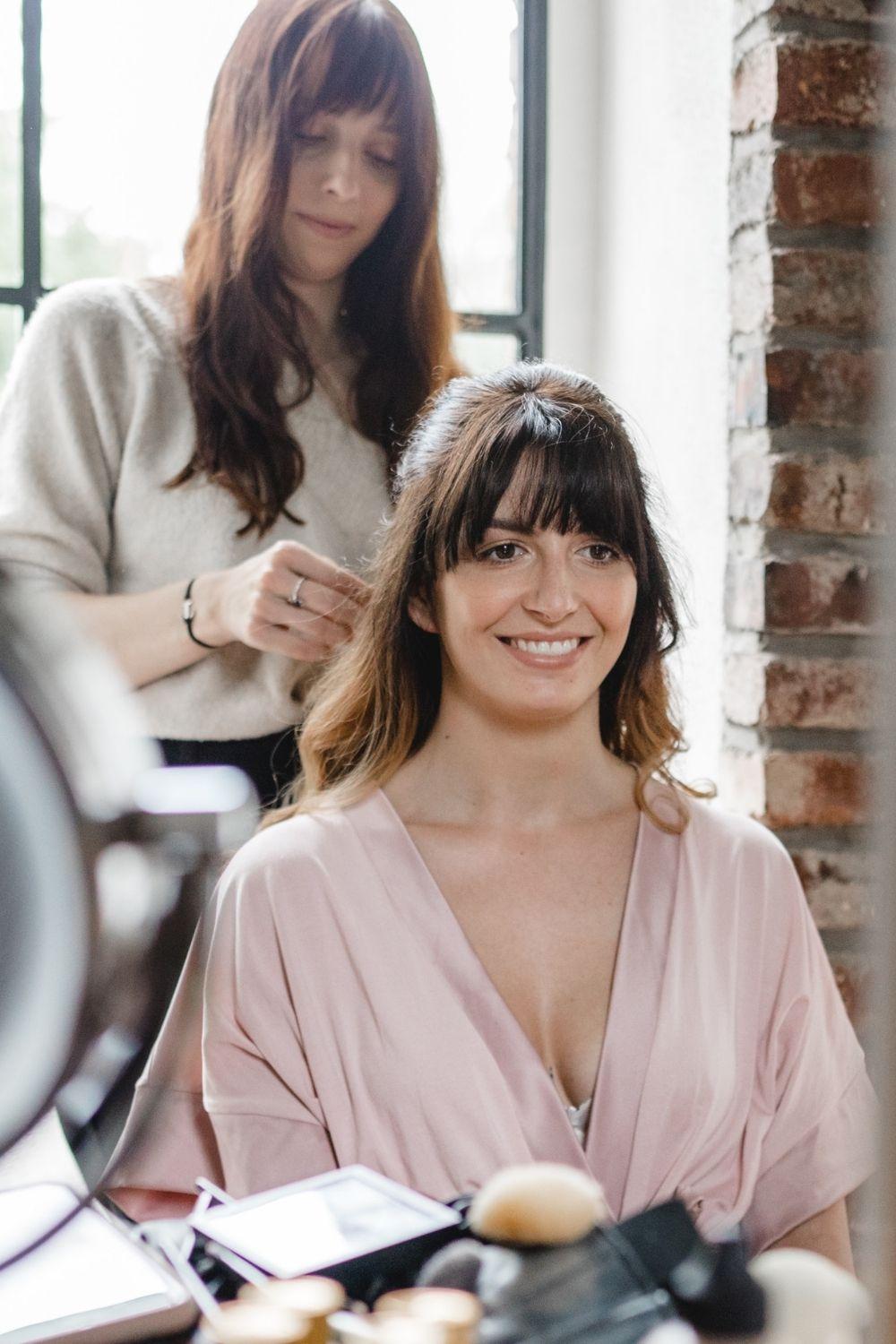 Lächelnde Braut im rosa Morgenmantel beim Hairstyling vor dem Spiegel; stimmungsvolle Vorbereitung auf die Hochzeit mit Fokus auf die Braut-Accessoires.