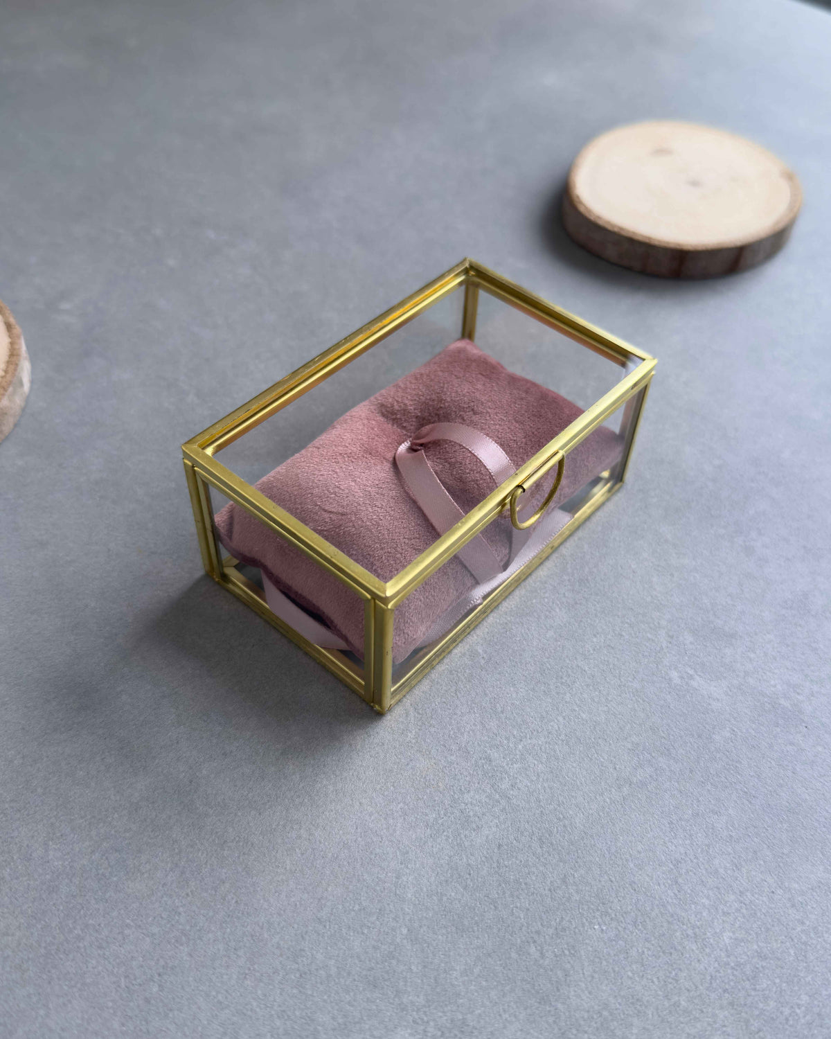 Geschlossenes Ringkästchen aus Glas mit goldenem Rahmen und altrosafarbenem Ringkissen mit Satinbandschlaufe in seitlicher Ansicht.