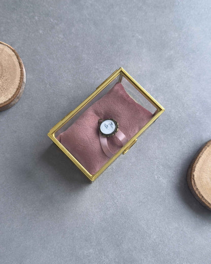 Geschlossenes Ringkästchen aus Glas mit goldenem Rahmen, altrosafarbenem Ringkissen und personalisiertem Anhänger in der Draufsicht.