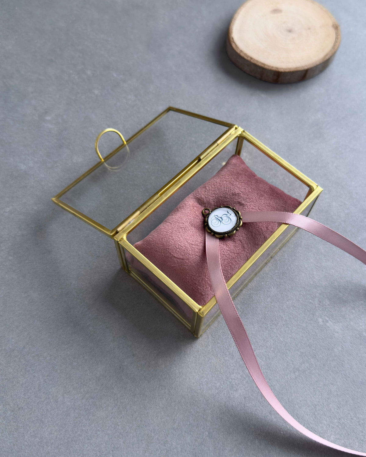 Offenes Ringkästchen aus Glas mit altrosafarbenem Ringkissen und personalisiertem Anhänger in schräger Ansicht.