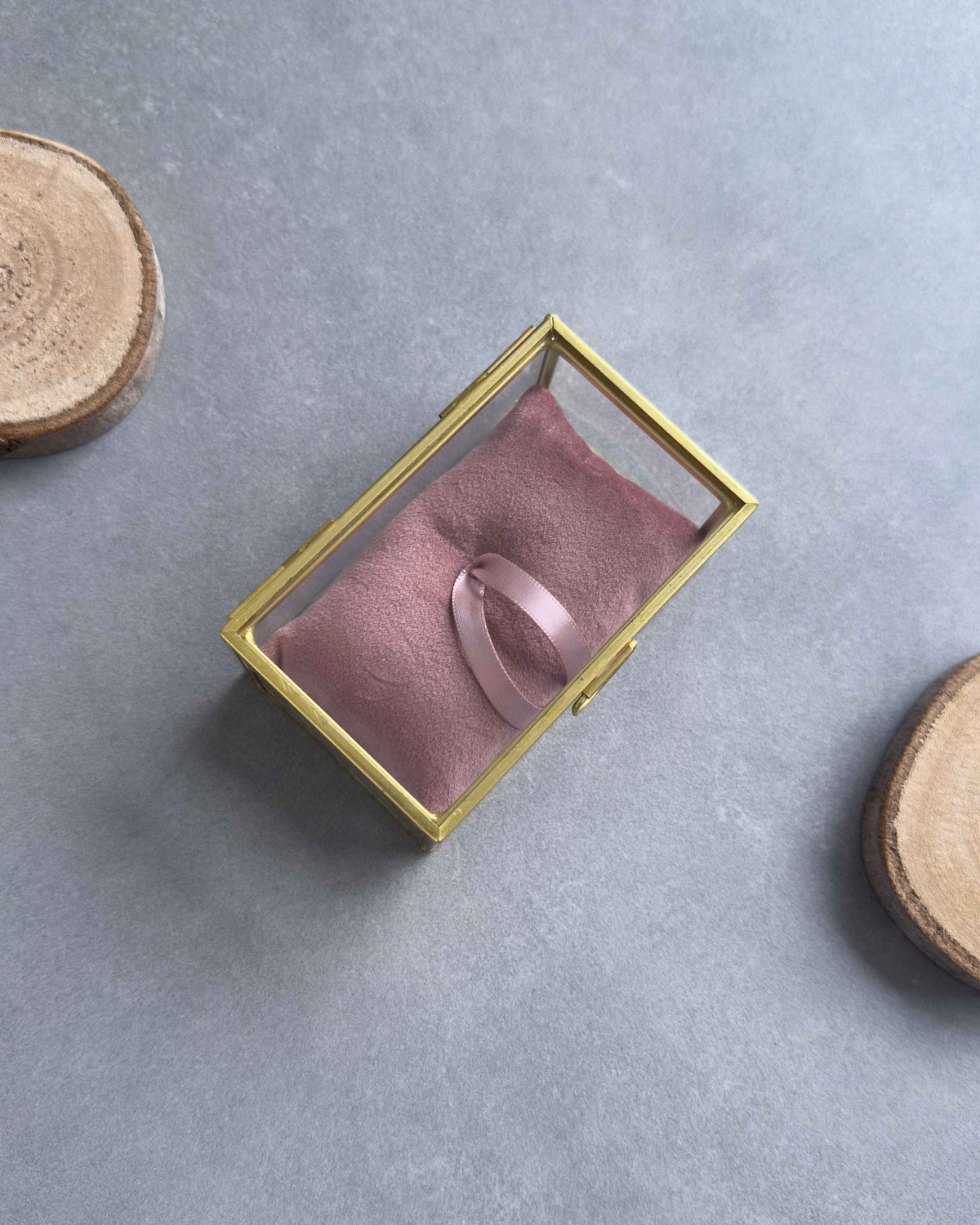 Geschlossenes Ringkästchen aus Glas mit goldenem Rahmen und altrosafarbenem Ringkissen mit Satinbandschlaufe, von oben fotografiert.