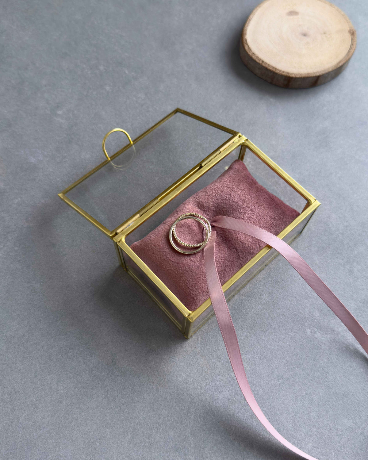 Offenes Ringkästchen aus Glas mit goldenem Rahmen, Ringkissen in Altrosa und zwei Eheringen in schräger Ansicht.