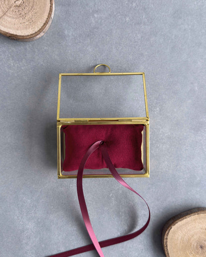 Offenes Ringkästchen aus Glas mit goldfarbenem Rahmen und rotem Ringkissen mit Satinband in der Frontansicht.