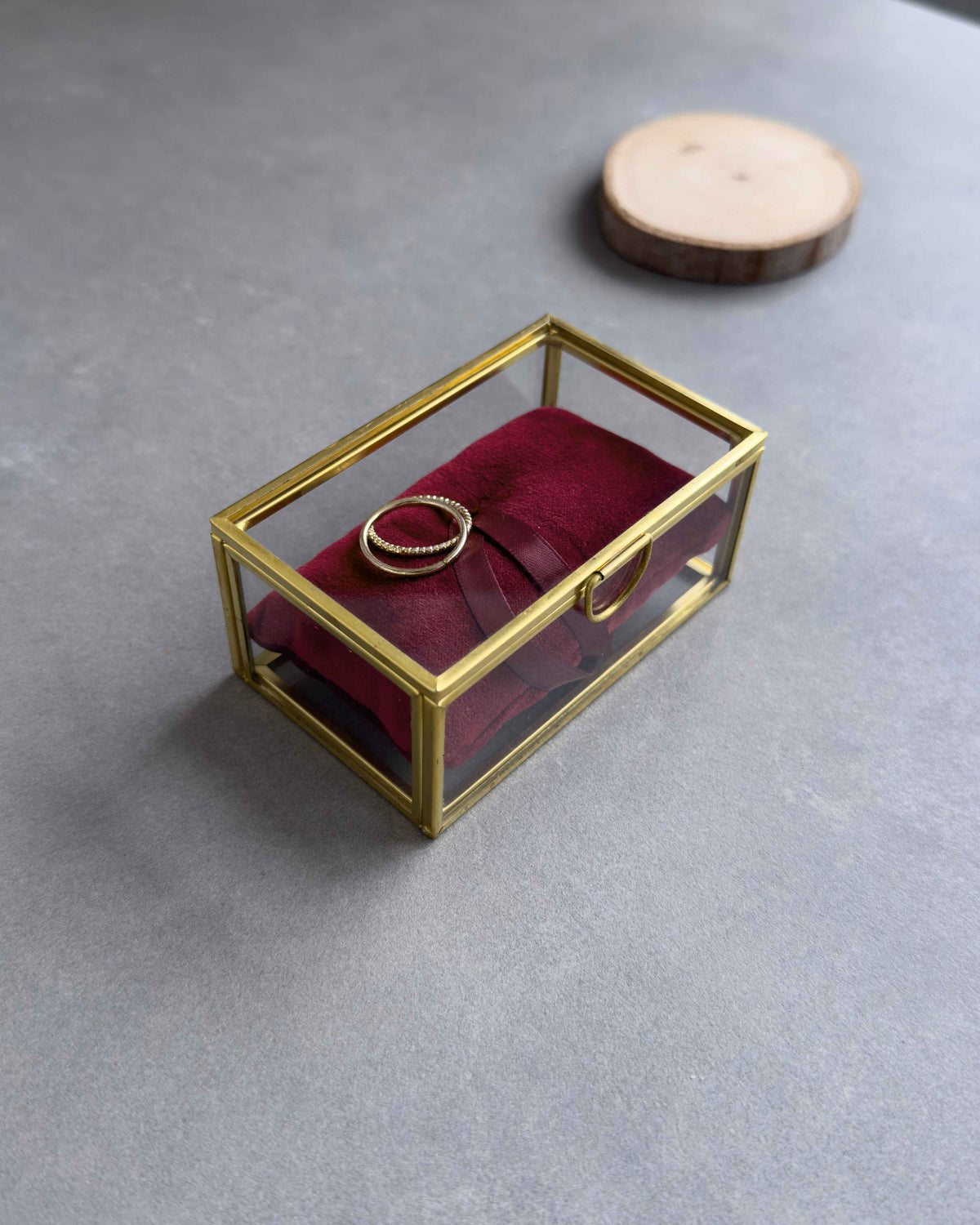 Geschlossenes Ringkästchen aus Glas mit goldenem Rahmen, rotem Ringkissen und Eheringen in seitlicher Ansicht.