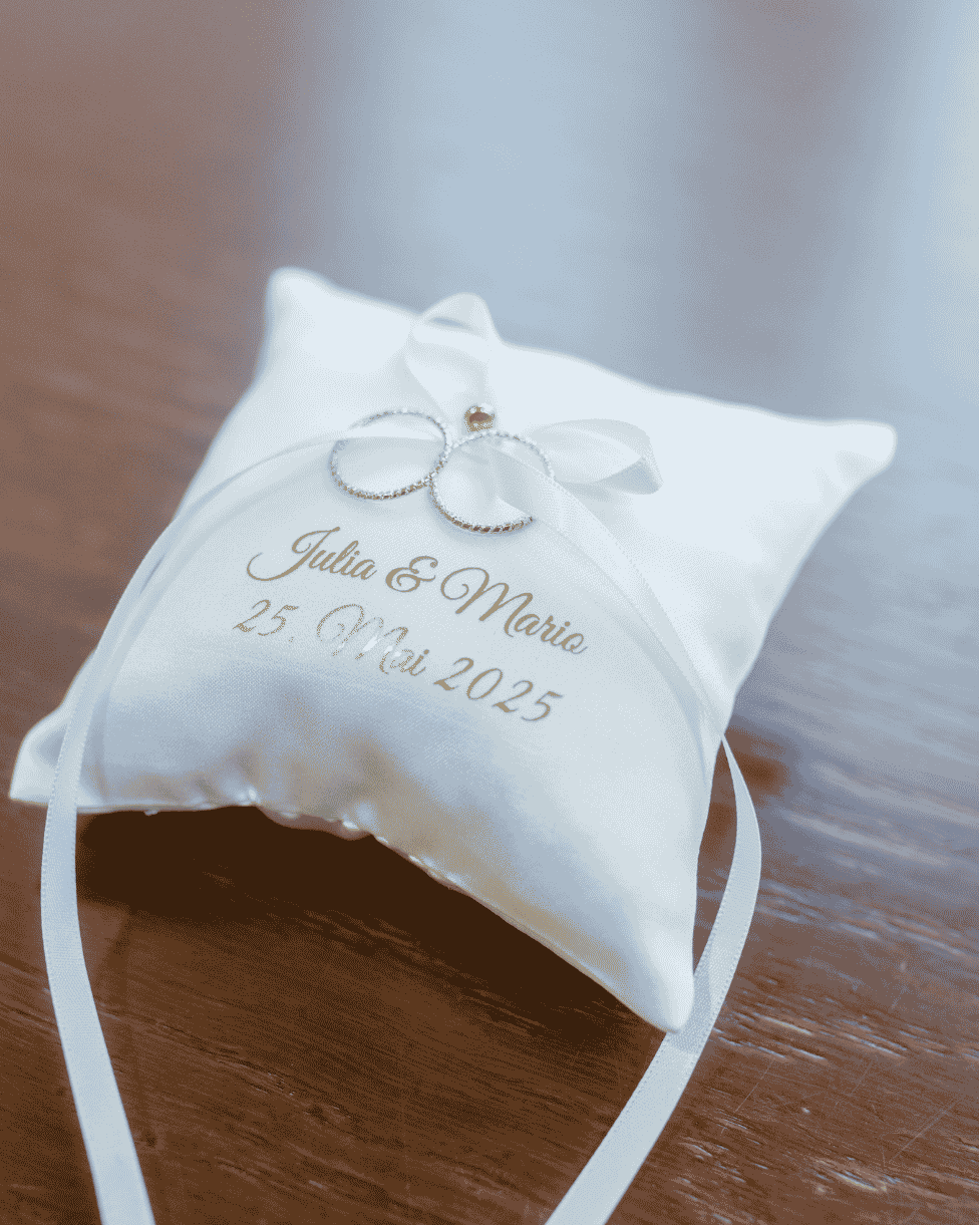 Klassisches weißes Ringkissen von Nadelspitze mit goldener Personalisierung „Julia & Mario“, Hochzeitsdatum und zwei Eheringen auf einem Holztisch.
